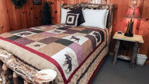 A wooden-paneled room with a bed featuring a quilt with animal prints, a nightstand with a lamp, and a wall-mounted decoration. - Lawn Lane A (Estes Park)