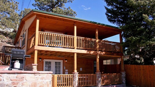 A two-story wooden cabin with a balcony, stone foundation, and a wooden fence. - Lawn Lane A (Estes Park)