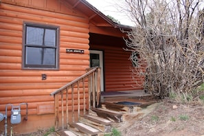 A log cabin with a wooden staircase leading to a front door, a sign reading "Blue Spruce B", and a small porch area.
