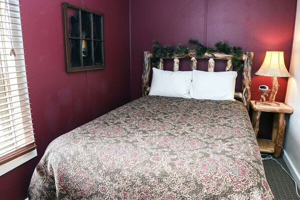A bedroom with a bed, a window with blinds, a wooden headboard with greenery, and a bedside lamp. - Lazy R Cottages: 6 (Estes Park)
