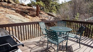 Eine Holzterrasse mit rundem Tisch und Stühlen, einem Grill und Blick auf einen bewaldeten, felsigen Hügel.