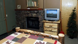 A room with a stone fireplace, a television on a wooden stand, a Christmas tree, and a quilt on the bed. - Timber Creek Chalets: 2B (Estes Park)