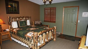 A bedroom with a rustic wooden bed frame, a green wall, a window with blinds, and a door with a welcome sign. - Timber Creek Chalets: 2B (Estes Park)