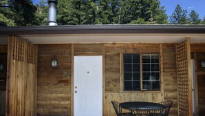 A wooden cabin with a white door, a window with a grid pattern, and a patio with a table and chairs. - Timber Creek Chalets: 2B (Estes Park)