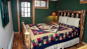 A bedroom with a wooden headboard, a quilted bedspread, a nightstand with a lamp, and a window with blinds.