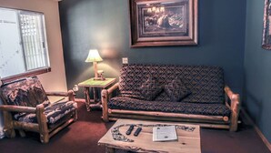 Un salon chaleureux avec un canapé à motifs, un fauteuil en bois, une petite table avec des télécommandes et un livre, et un tableau encadré au mur.