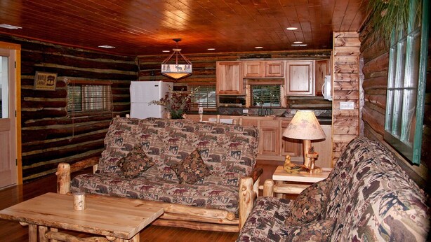 A log cabin interior with a wooden ceiling, a patterned sofa, a wooden coffee table, a kitchen area with wooden cabinets, and a stone wall with a window. - Triple R Cottages: 8 (Estes Park)
