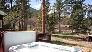 A hot tub in a wooded backyard with a wooden deck and a small house in the background. - Triple R Cottages: 8 (Estes Park)