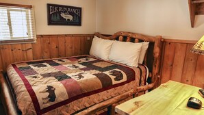 A wooden bed with a quilt featuring elk and deer, a wooden headboard, and a small table with a lamp and mouse. - Triple R Cottages: 3 (Estes Park)
