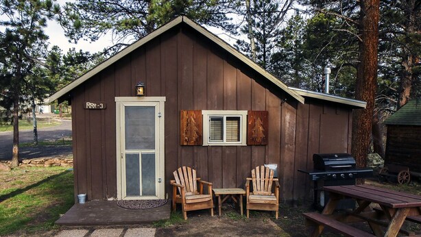 A small wooden cabin with a door, two Adirondack chairs, a picnic table, and a grill. - Triple R Cottages: 3 (Estes Park)