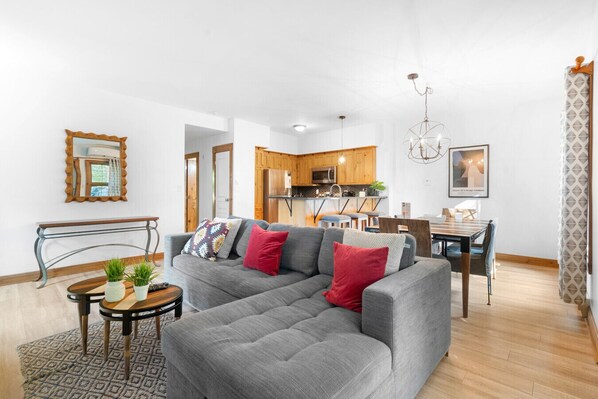 Een moderne woonkamer met een grijze modulaire bank, een klein rond tafeltje met planten en een kamerbreed tapijt. Achterin is een houten bankje en een eettafel met stoelen te zien.