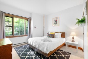 Een hotelkamer met een groot bed, een nachtkastje met een lamp, een raam met gordijnen, een kunstwerk in een lijst aan de muur en een hangplant.