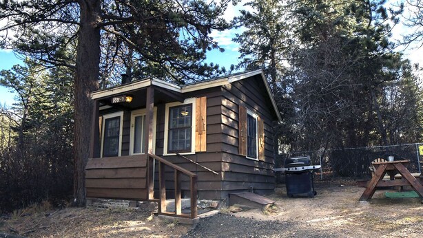A small cabin with a wooden deck and a chimney, surrounded by trees. - Triple R Cottages: 7 (Estes Park)