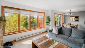 Ett modernt vardagsrum med stora fönster, en grå soffa, ett soffbord i trä och en matplats i bakgrunden.