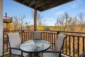 A covered outdoor deck with a table and chairs, overlooking a forested area with autumn foliage.