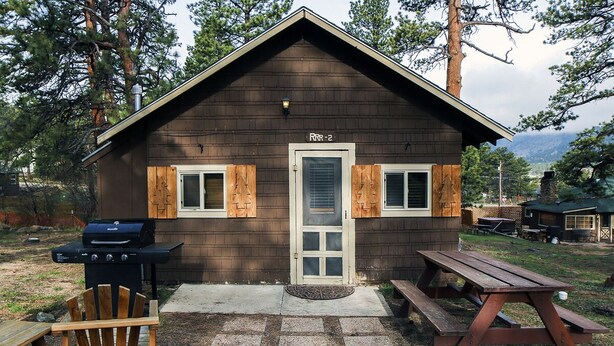 A rustic cabin with a wooden exterior, a grill, a picnic table, and a chair. - Triple R Cottages: 2 (Estes Park)
