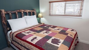 A bed with a wooden headboard, a quilt with animal prints, a bedside lamp, and a window with blinds. - Triple R Cottages: 2 (Estes Park)