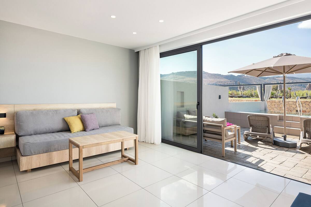 A modern living room with a sofa, a wooden coffee table, and a glass door leading to an outdoor patio with a table, chairs, and an umbrella.