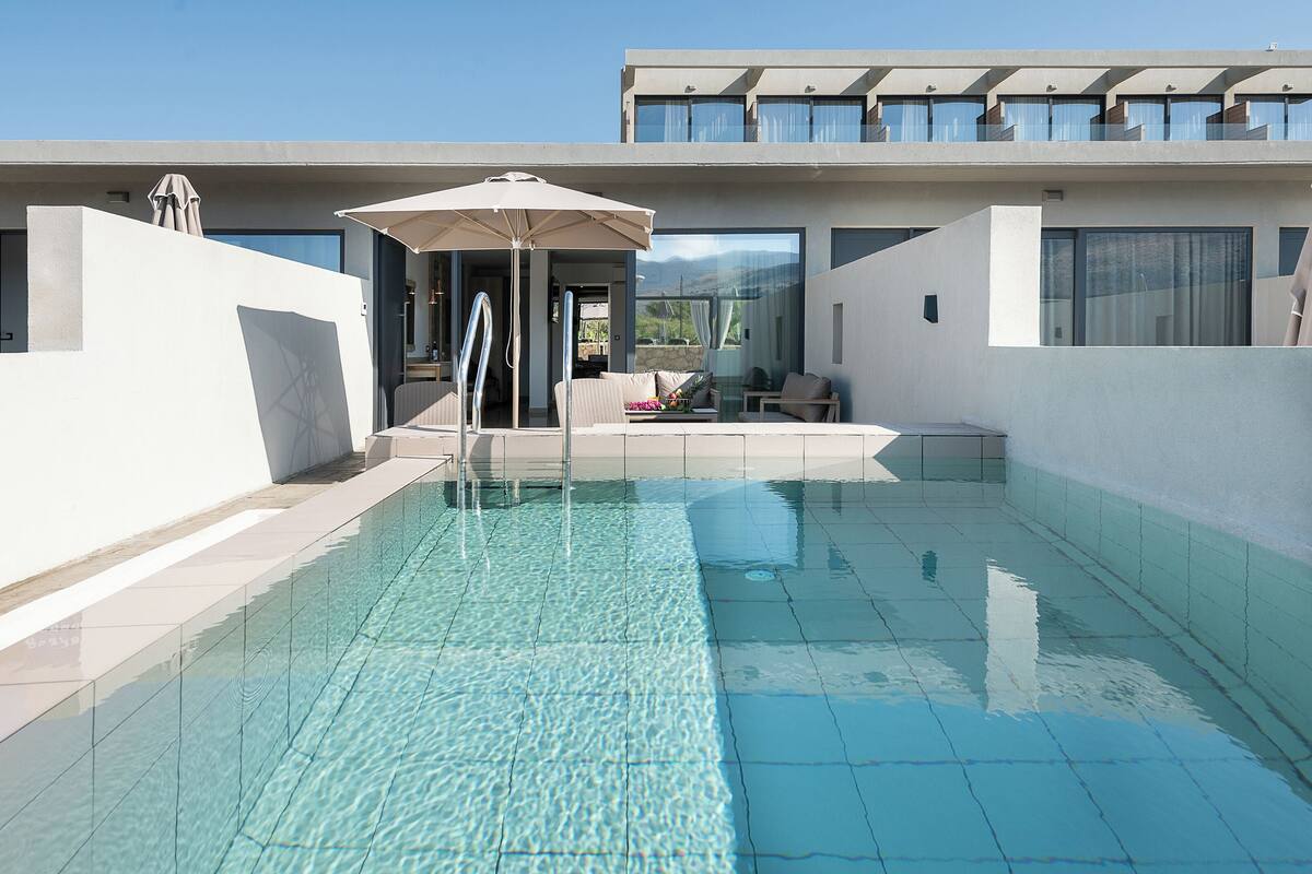 A modern outdoor swimming pool with a clear blue water surface, a lounge area with seating, and a contemporary building with large windows and balconies.
