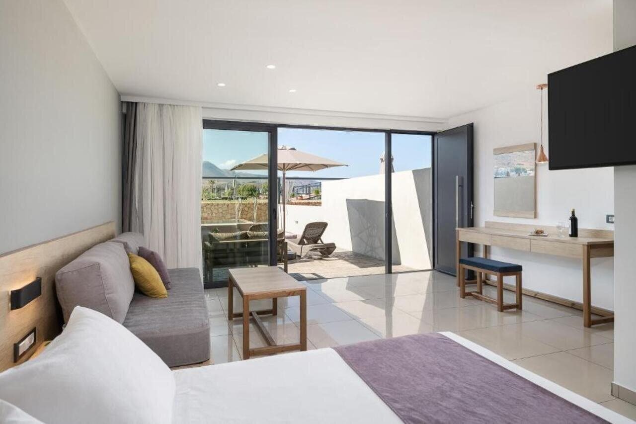 Habitación de hotel moderna con un amplio ventanal corredero que da a un patio, un sofá, una mesita, un banco y un televisor montado en la pared.