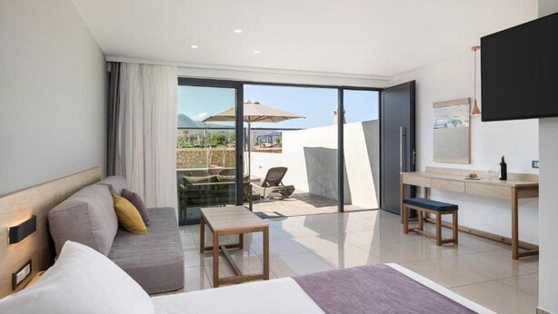 A modern hotel room with a large sliding glass door leading to a patio area, a sofa, a small table, a bench, and a TV mounted on the wall. - Chq Galini Palace (Platanias)