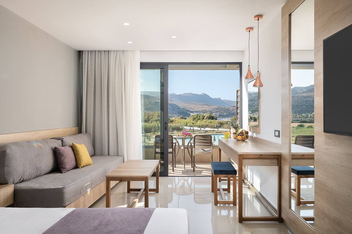 A modern hotel room with a balcony, a sofa, a dining table, and a view of the mountains.