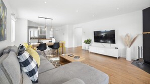 Ein modernes Wohnzimmer mit einem grauen Sofa, einem Essbereich mit Holztisch und -stühlen, einer Küche im Hintergrund und einem großen Fernseher an der Wand.