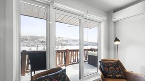 Ein Zimmer mit großem Fenster und Blick auf eine verschneite Landschaft, eine Holzterrasse davor sowie zwei Ledersessel mit gemusterten Kissen.