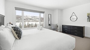 Ein Schlafzimmer mit einem großen Bett, einem Schminktisch, einem Sessel und Blick auf eine verschneite Landschaft durch ein großes Fenster.