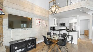 A modern living room with a large flat-screen TV, a glass dining table with black chairs, a wooden entertainment unit, and a kitchen area with white cabinets and a microwave.