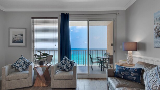 Ein Wohnzimmer mit einer Couch, zwei Sesseln, einem Couchtisch und einem Esstisch mit Stühlen. Ein großes Fenster bietet Meerblick und es gibt einen Balkon.