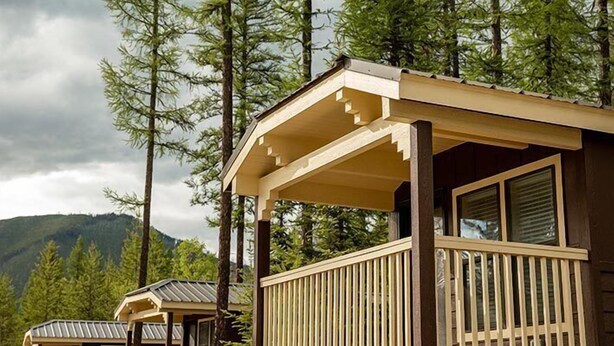 A cabin with a wooden deck and large windows, surrounded by tall pine trees and a mountain in the background. - West Glacier Cabin Village (West Glacier)