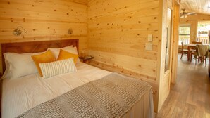 A wooden cabin bedroom with a bed, pillows, a bedside table, and a view of the dining area. - West Glacier Cabin Village (West Glacier)