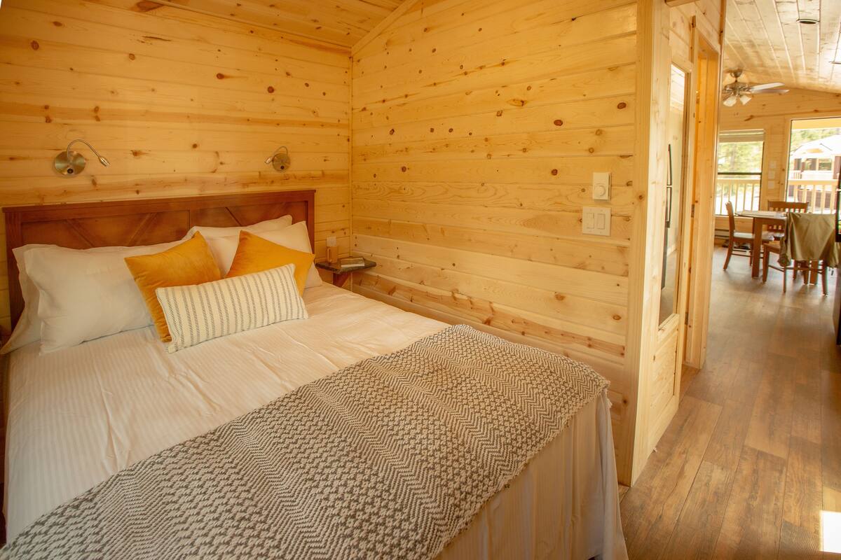 A wooden cabin bedroom with a bed, pillows, a bedside table, and a view of the dining area.