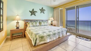 Une chambre avec un lit, deux tables de chevet, un ventilateur de plafond et une vue sur l’océan, visible à travers une porte-fenêtre coulissante.