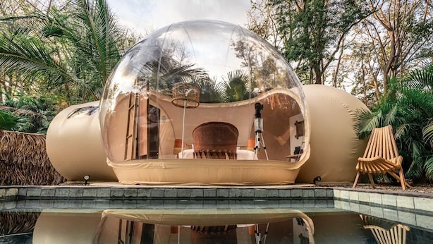 Una cúpula transparent sobre una piscina, amb una zona d'seients a l'interior, envoltada de vegetació.
