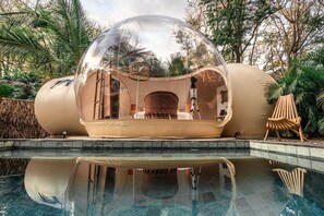 A transparent dome structure over a pool with a seating area inside, surrounded by greenery. - Satori The Bubble Experience Lodge (Nosara)