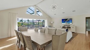 A spacious dining area with a large table and chairs, a modern chandelier, and a view of the outdoors. - Elegance Redefined, Simply Seconds Away from the Beach! (Marco Island)