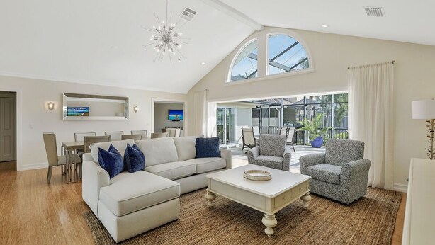 A spacious living room with a large sectional sofa, a coffee table, and patterned armchairs. A television is mounted on the wall, and there is a dining area with a table and chairs. - Elegance Redefined, Simply Seconds Away from the Beach! (Marco Island)