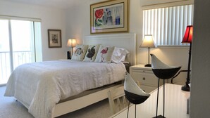 A bedroom with a bed, bedside lamps, a ceiling fan, a nightstand, a desk with a chair, and a window with blinds. - CP 421 2 Bedroom Ocean View Condo - Paradise Vacation Rental in Ocean Village (Fort Pierce)
