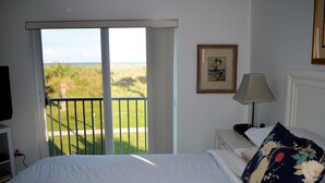 A bedroom with a large bed, a nightstand, a lamp, a framed picture, and a view of a grassy area through a sliding door. - CP 421 2 Bedroom Ocean View Condo - Paradise Vacation Rental in Ocean Village (Fort Pierce)