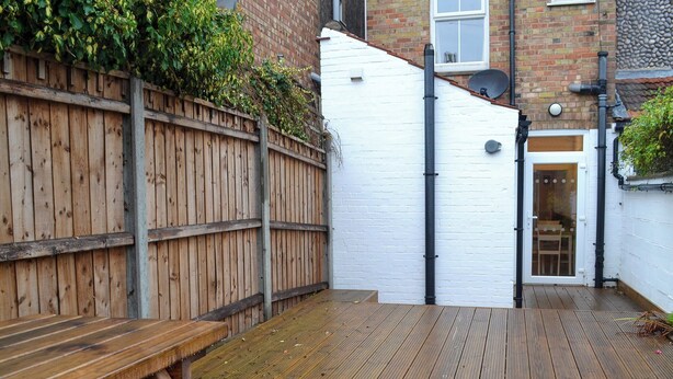 Une terrasse en bois avec un banc, un mur en briques blanches et une porte donnant sur une pièce.