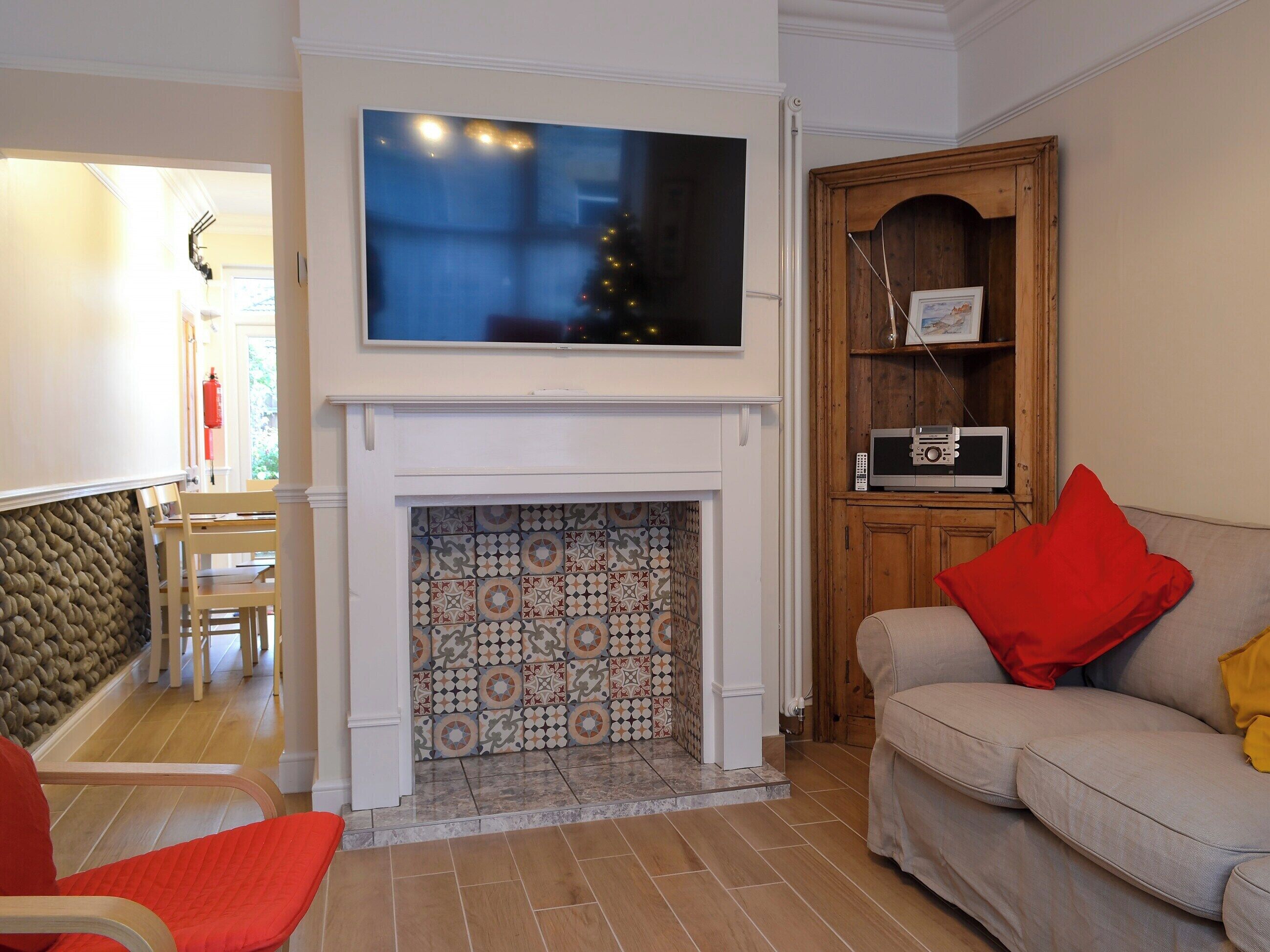 A living room with a fireplace, a television mounted above it, a sofa with red and yellow cushions, and a wooden cabinet with a radio.