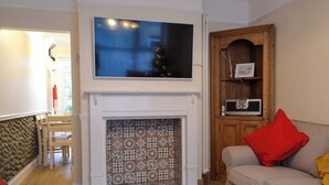 Un salon doté d’une cheminée, avec un téléviseur fixé au-dessus, un canapé orné de coussins rouges et jaunes, et un meuble en bois abritant une radio.