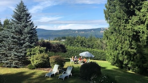 Un jardín con dos sillones de descanso, una mesita y un grupo de personas bajo una sombrilla.
