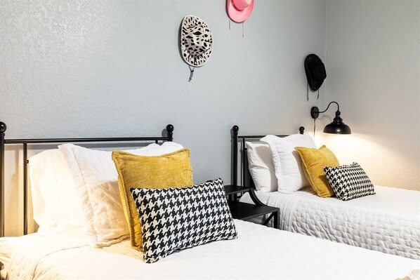 Dos camas individuales con ropa blanca y almohadas estampadas, una lámpara negra y una lámpara de pared.