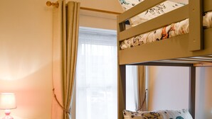 Een stapelbed met opvallende kussens, een nachtkastje met een lamp en een raam met gordijnen.