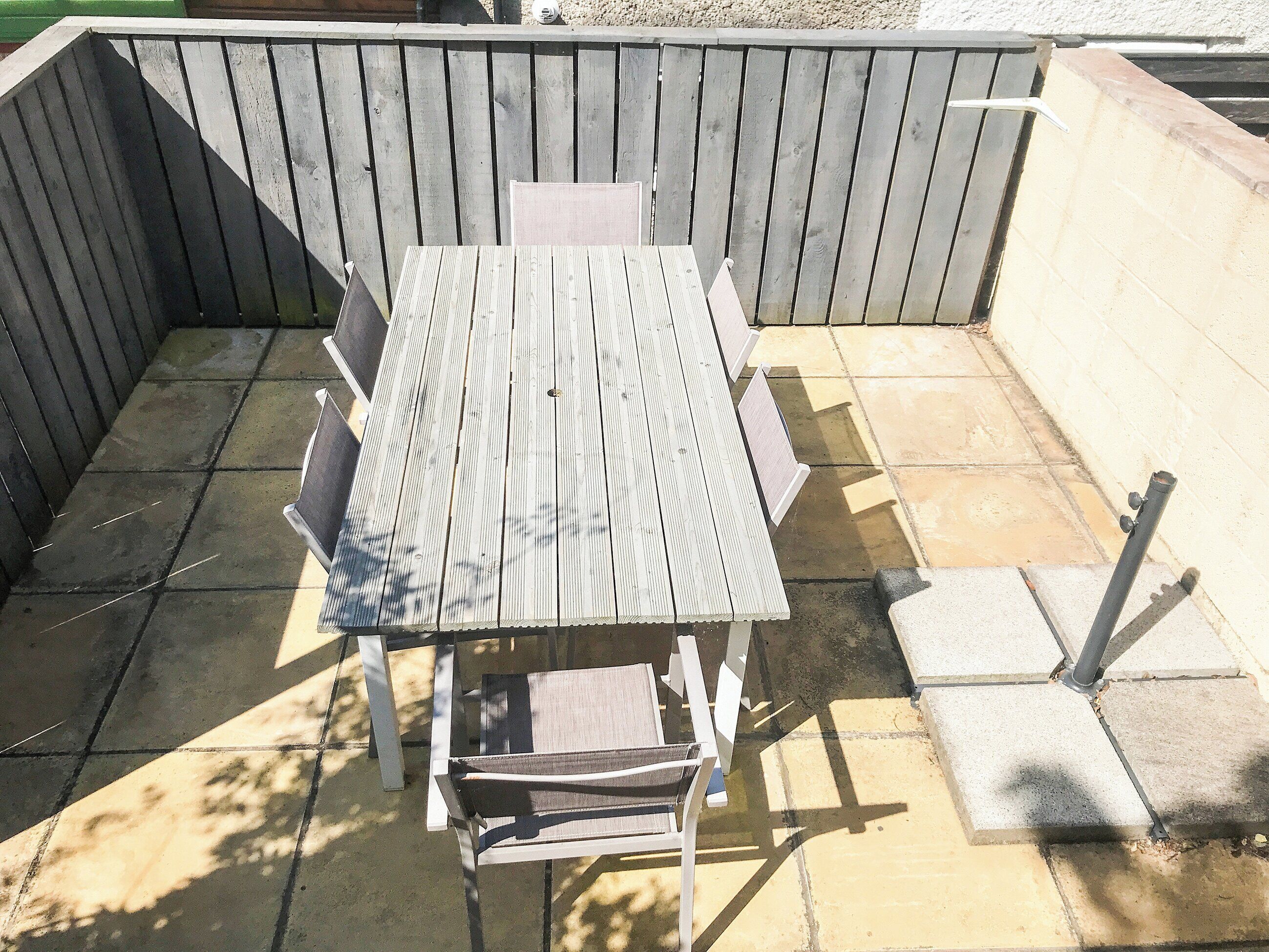 Mesa de madera con sillas en un patio exterior con suelo de baldosas y cerca de madera.