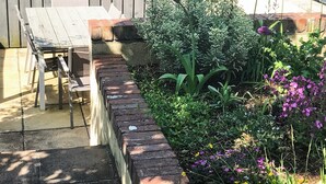 Een terras met een bakkenplanter van baksteen, tuinmeubilair en diverse planten en bloemen.