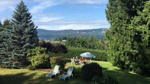 Un jardín con dos sillones de descanso, una mesita y un grupo de personas bajo una sombrilla.